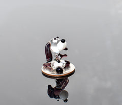 May include: A small glass figurine of a dog sitting on a silver coin. The dog is white with black eyes and a black nose. The dog's ears are brown and its tail is curled up. The dog is sitting on a silver coin with the words 'In God We Trust' and 'Liberty' visible.