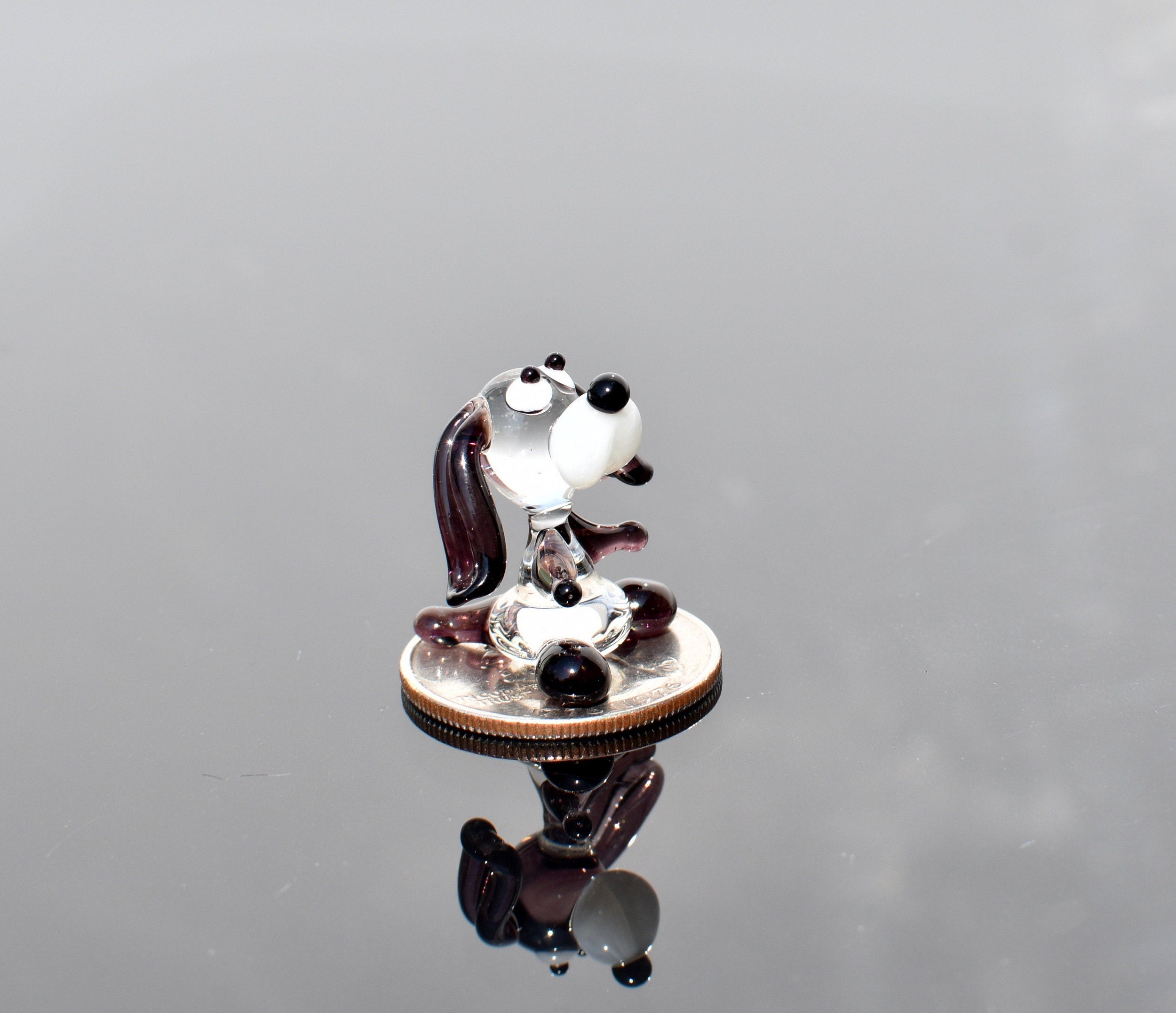 May include: A small glass figurine of a dog sitting on a silver coin. The dog is white with black eyes and a black nose. The dog's ears are brown and its tail is curled up. The dog is sitting on a silver coin with the words 'In God We Trust' and 'Liberty' visible.