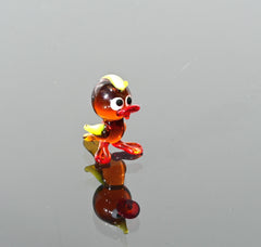 May include: A small glass duck figurine with a yellow beak, red feet, and brown body. The duck has large, black eyes and is standing on a reflective surface.