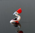 May include: A small glass figurine of a bird with a red head, white body, and black and purple accents. The bird is sitting on a reflective surface.