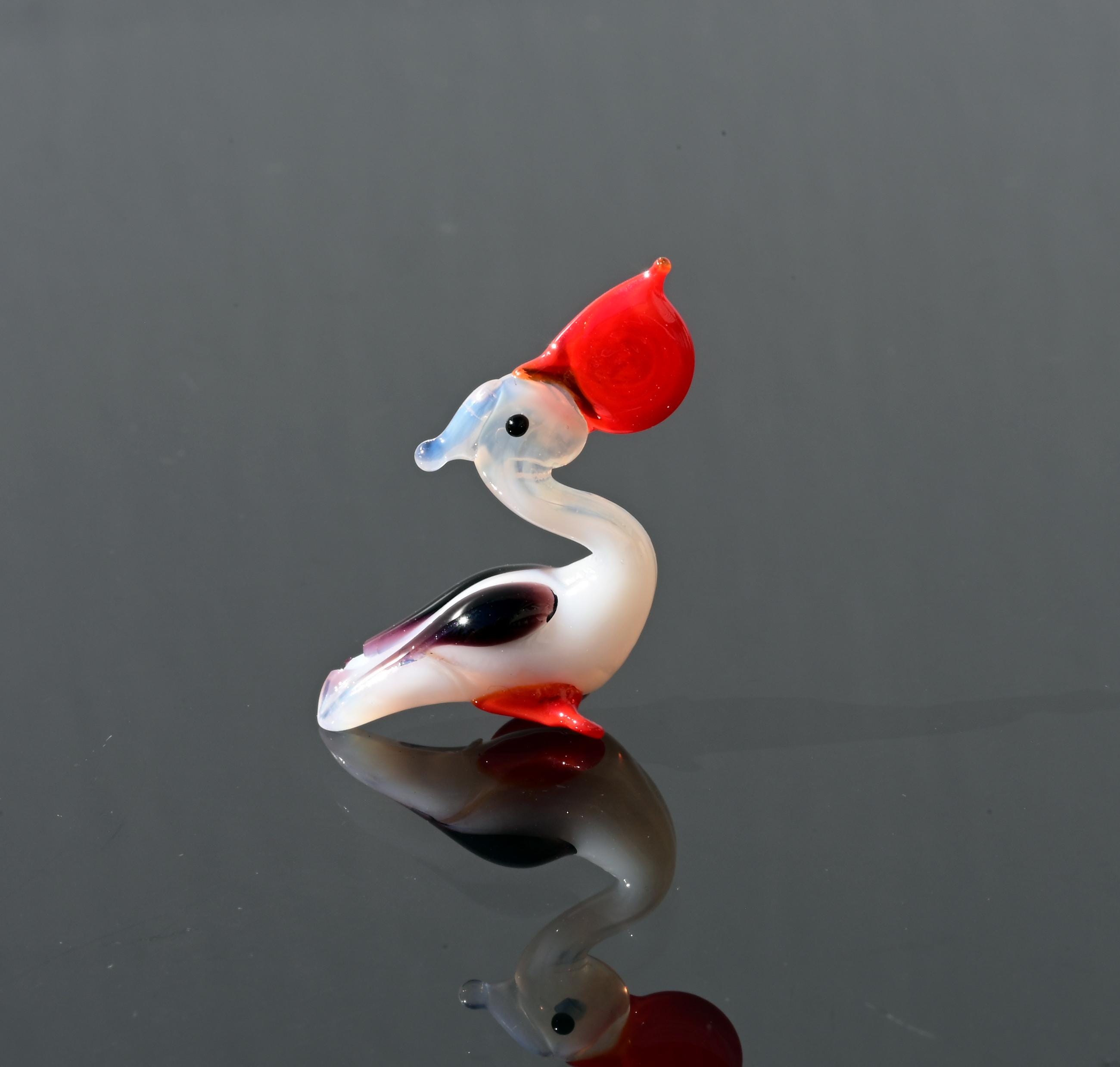 May include: A small glass figurine of a bird with a red head, white body, and black and purple accents. The bird is sitting on a reflective surface.
