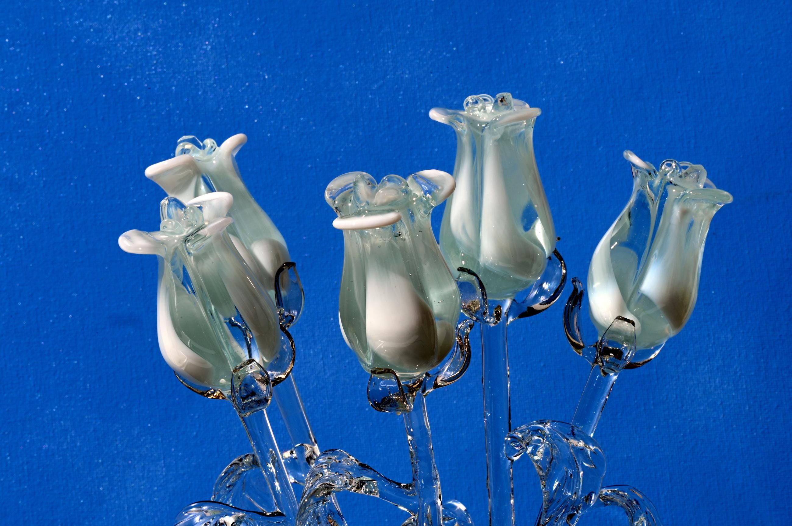 May include: A bouquet of five white glass roses with clear glass stems. The roses are arranged in a cluster and are set against a blue background.