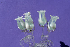 May include: A bouquet of five white glass roses with light blue accents. The roses are arranged in a vase and are on a purple background.