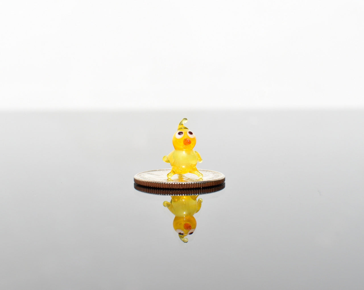 May include: A small yellow glass chick figurine with orange eyes and beak standing on a silver coin.