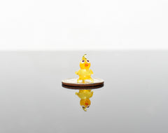 May include: A small yellow glass chick figurine with orange eyes and beak standing on a silver coin.