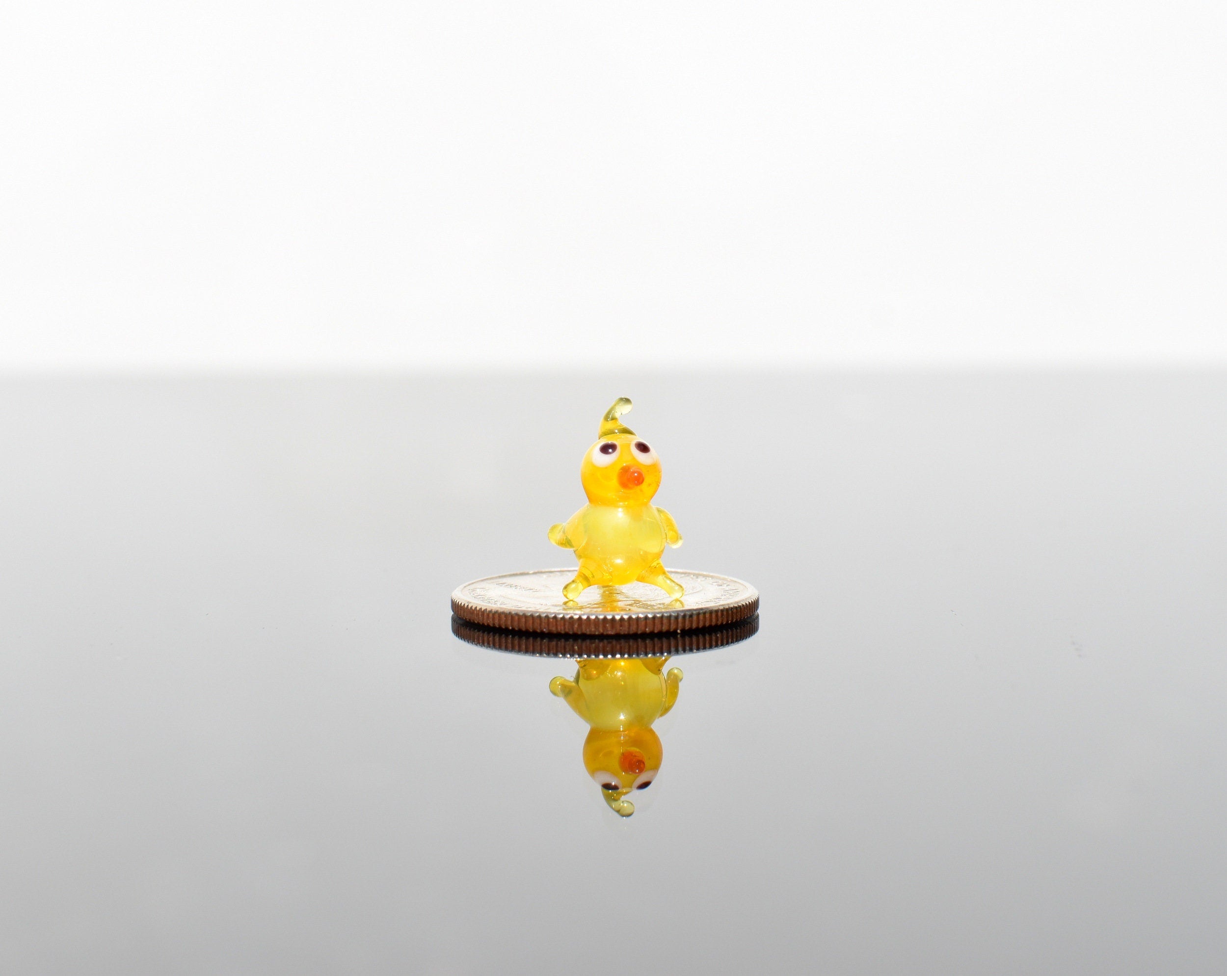May include: A small yellow glass chick figurine with orange eyes and beak standing on a silver coin.