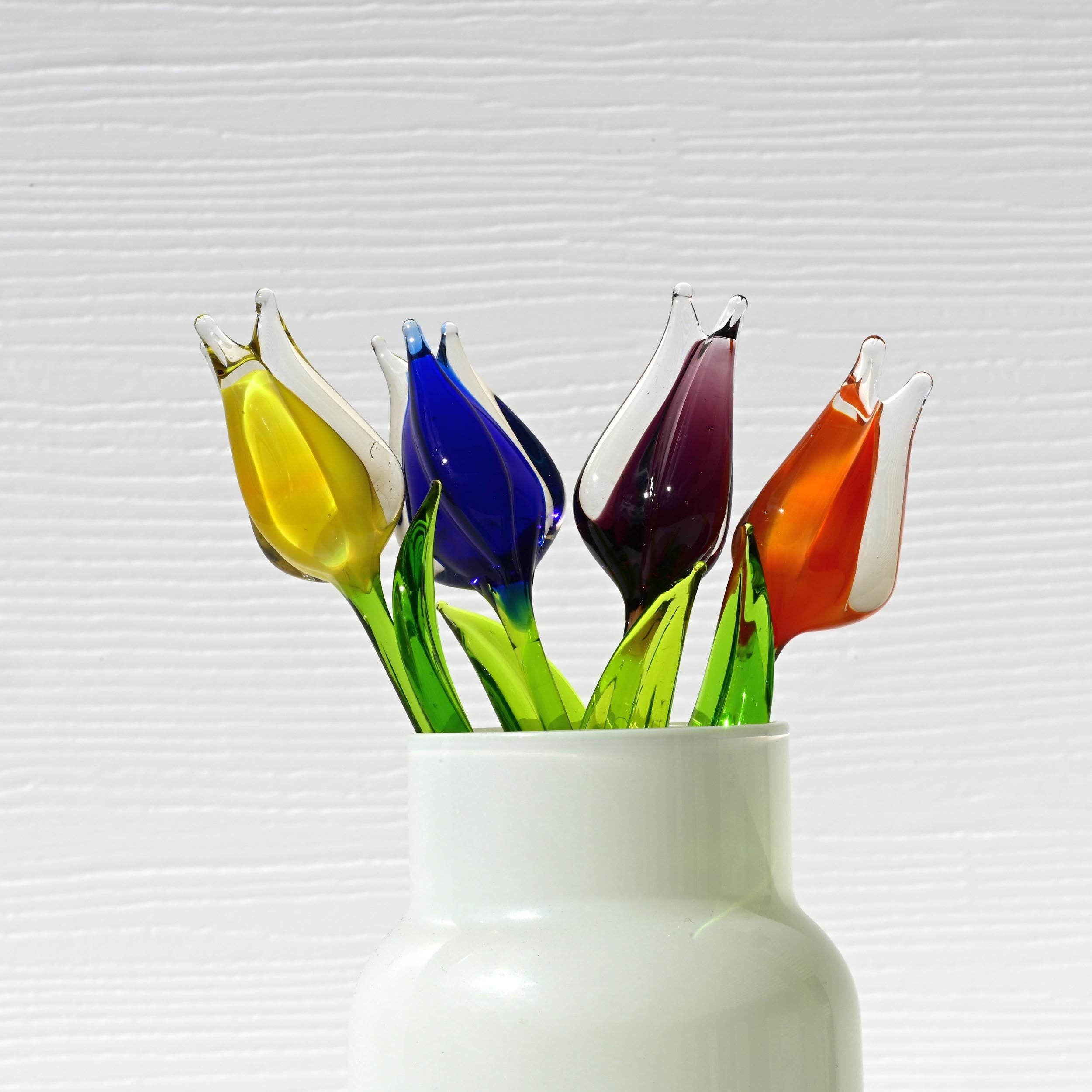 May include: Five glass tulips in a white vase.  The tulips are vibrant colors: yellow, blue, purple, and orange. The stems are green glass. The tulips are artfully arranged in a simple, elegant white vase.  These handcrafted glass flowers are a unique home decor accent.