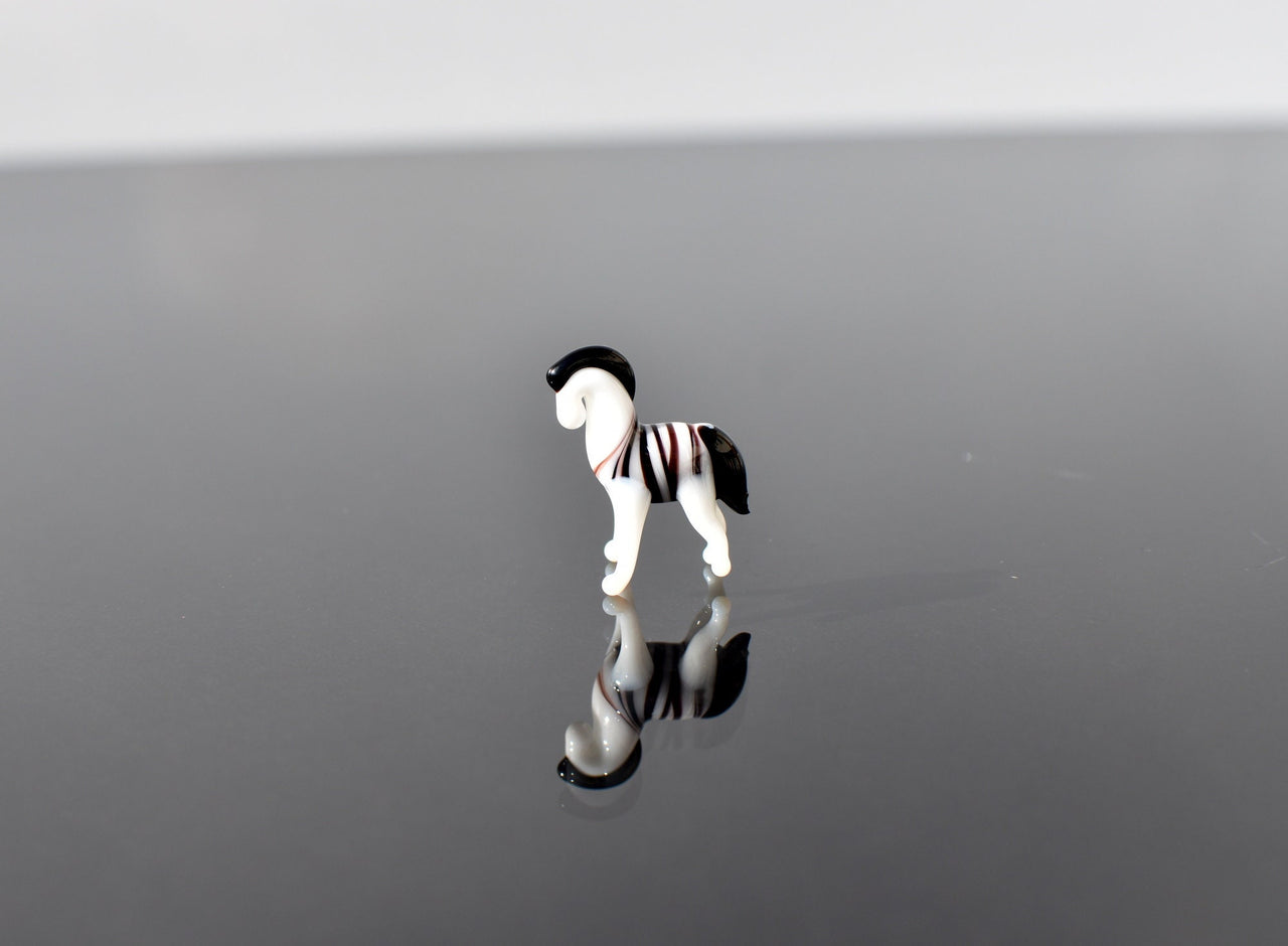 May include: A small glass zebra figurine with black and white stripes. The figurine is standing on a reflective surface.
