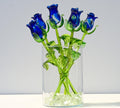 May include: A bouquet of five blue glass roses with green glass stems and leaves in a clear glass vase with white glass stones at the bottom